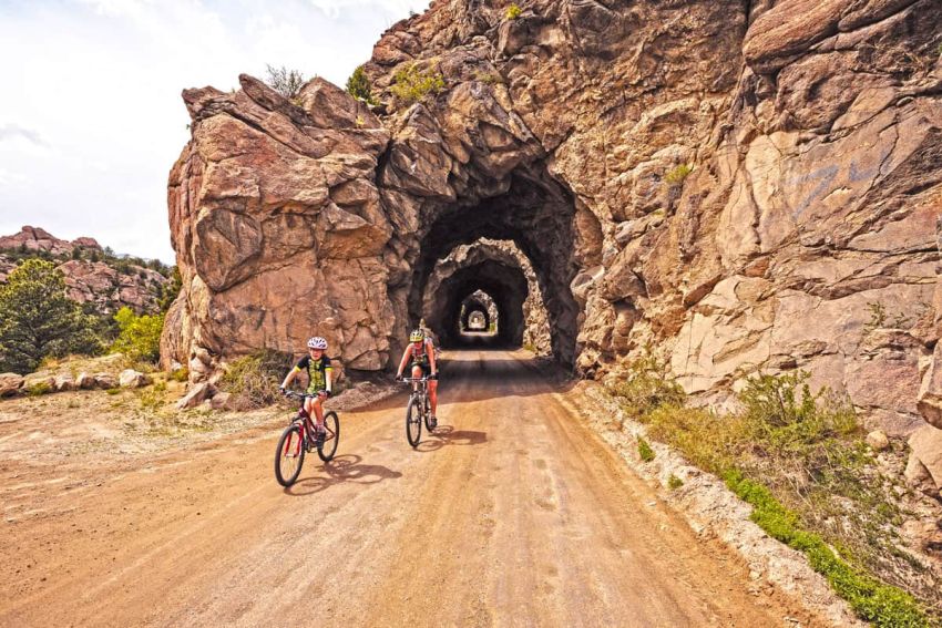 Two people biking in Colorado, down a long dirt road through consecutive tunnels cut into the rock.